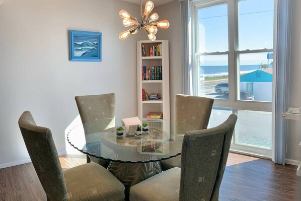 A dining area with a glass table surrounded by chairs, a bookshelf, and a view of the sea through a large window.