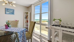 A room with a dining table, chairs, a bookshelf, a potted plant, and a view of the beach through large windows. - Pelican Suite | Bombora Suites | Steps To Flagler Beach & Restaurants (Flagler Beach)
