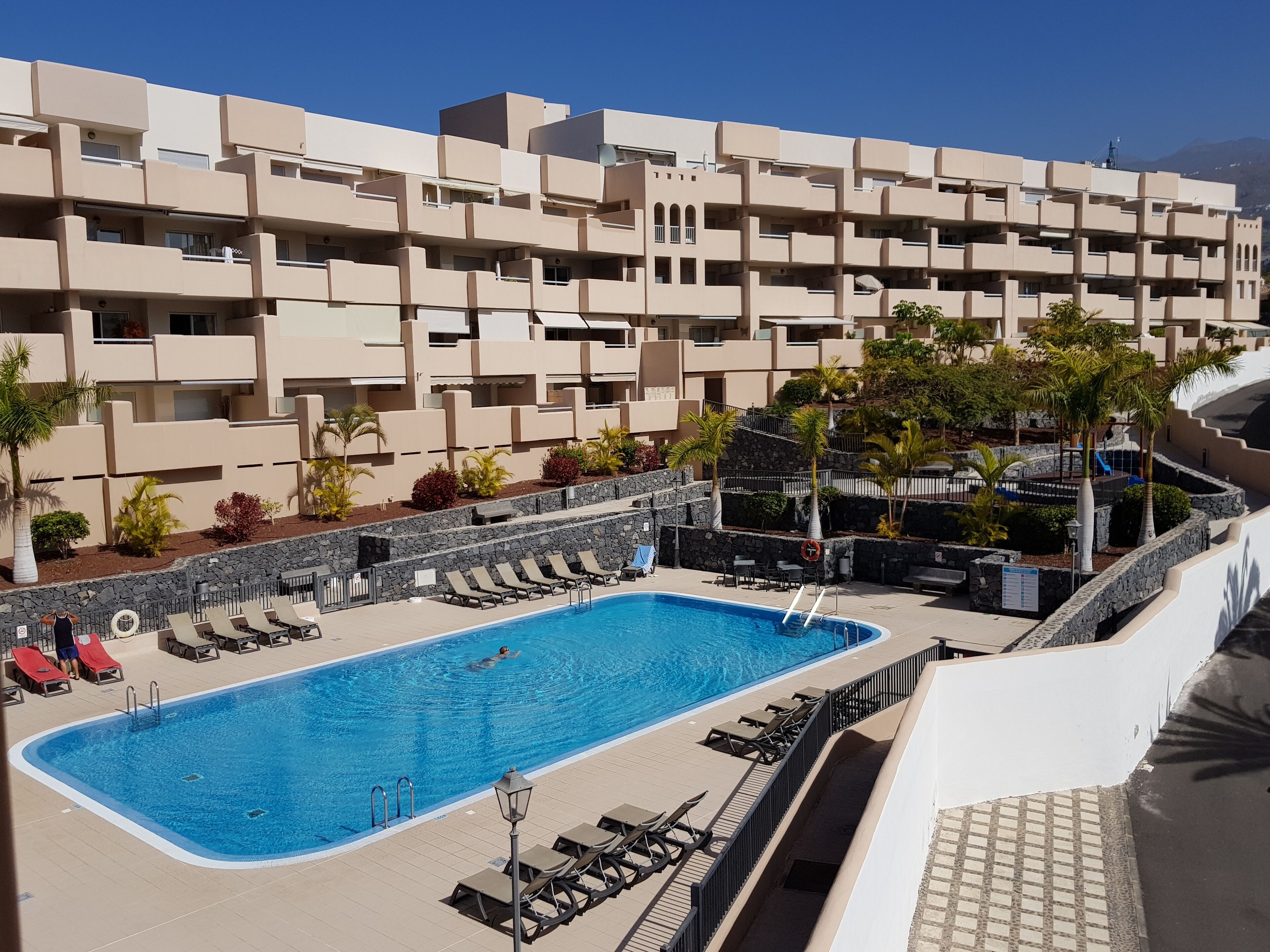 Un hotel con piscina, varios balcones y un área ajardinada con palmeras.