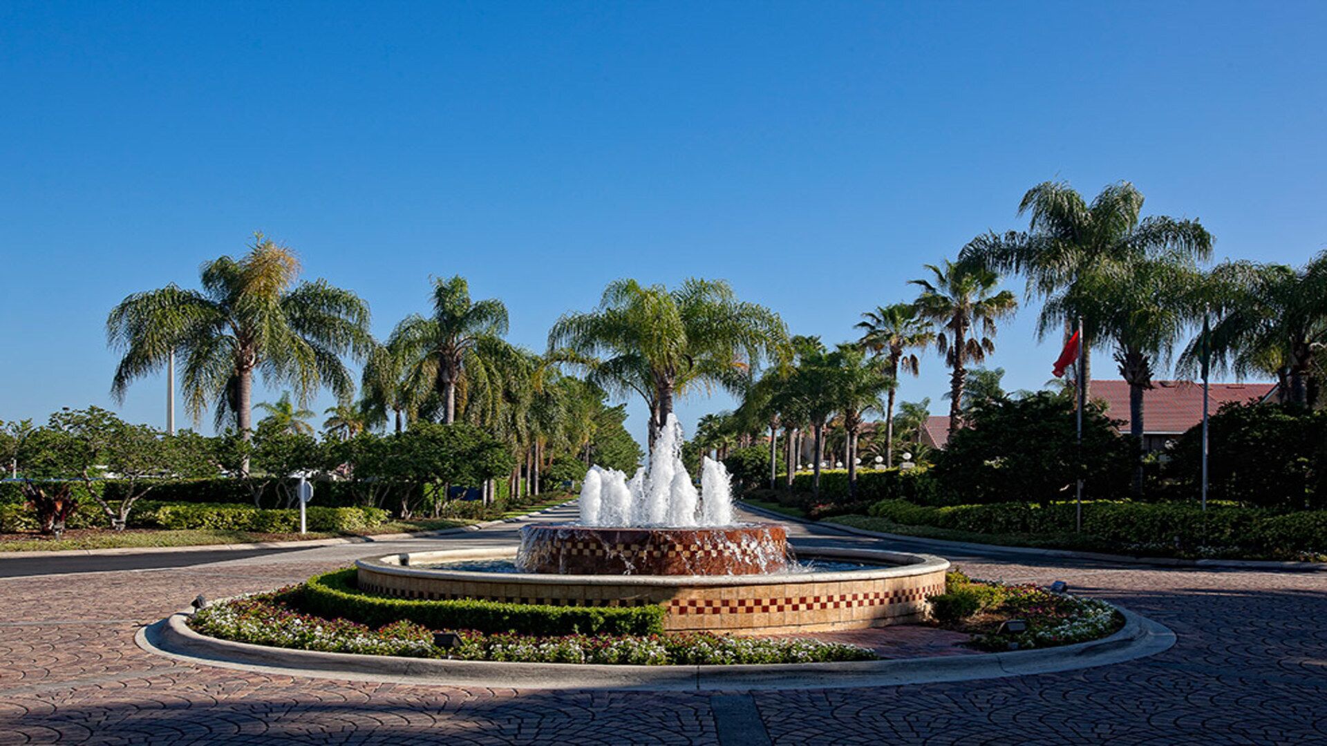 A circular fountain with water jets in the center, surrounded by a paved walkway and palm trees.