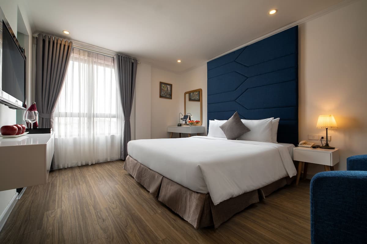 A hotel room with a large bed, a desk, a chair, a bedside table, a lamp, and a window with curtains.