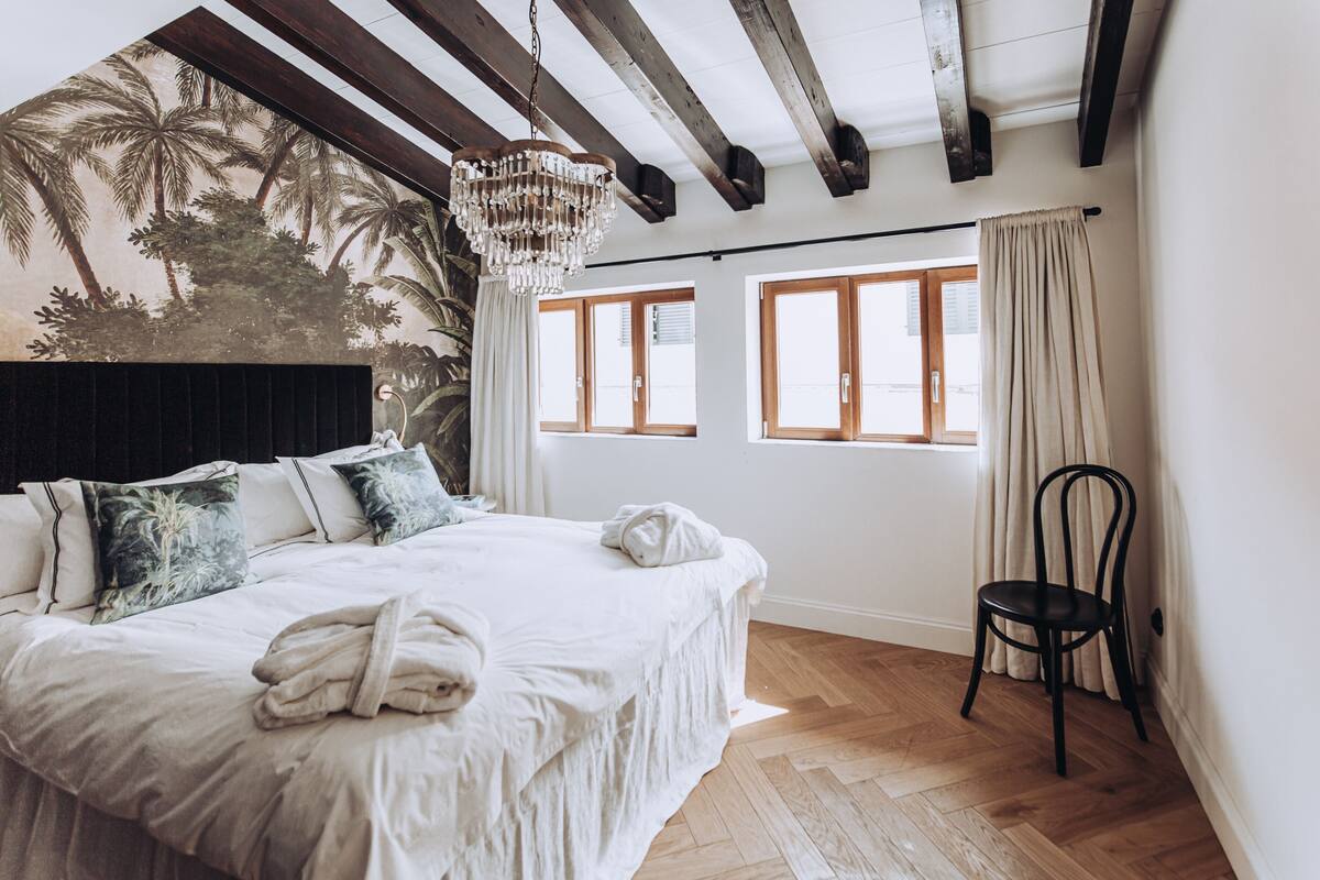 A bedroom with a bed, a chandelier, a chair, and a window with curtains.