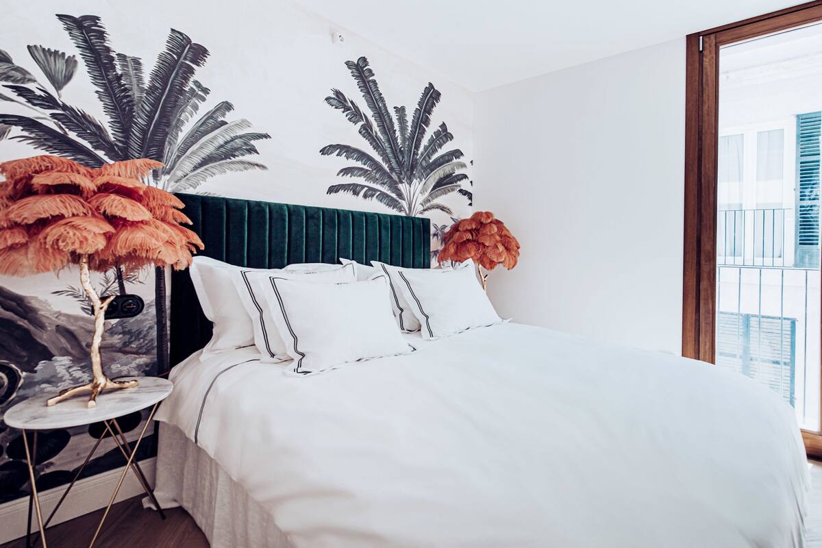 A bedroom with a large bed, a green headboard, and a wall mural of palm leaves.