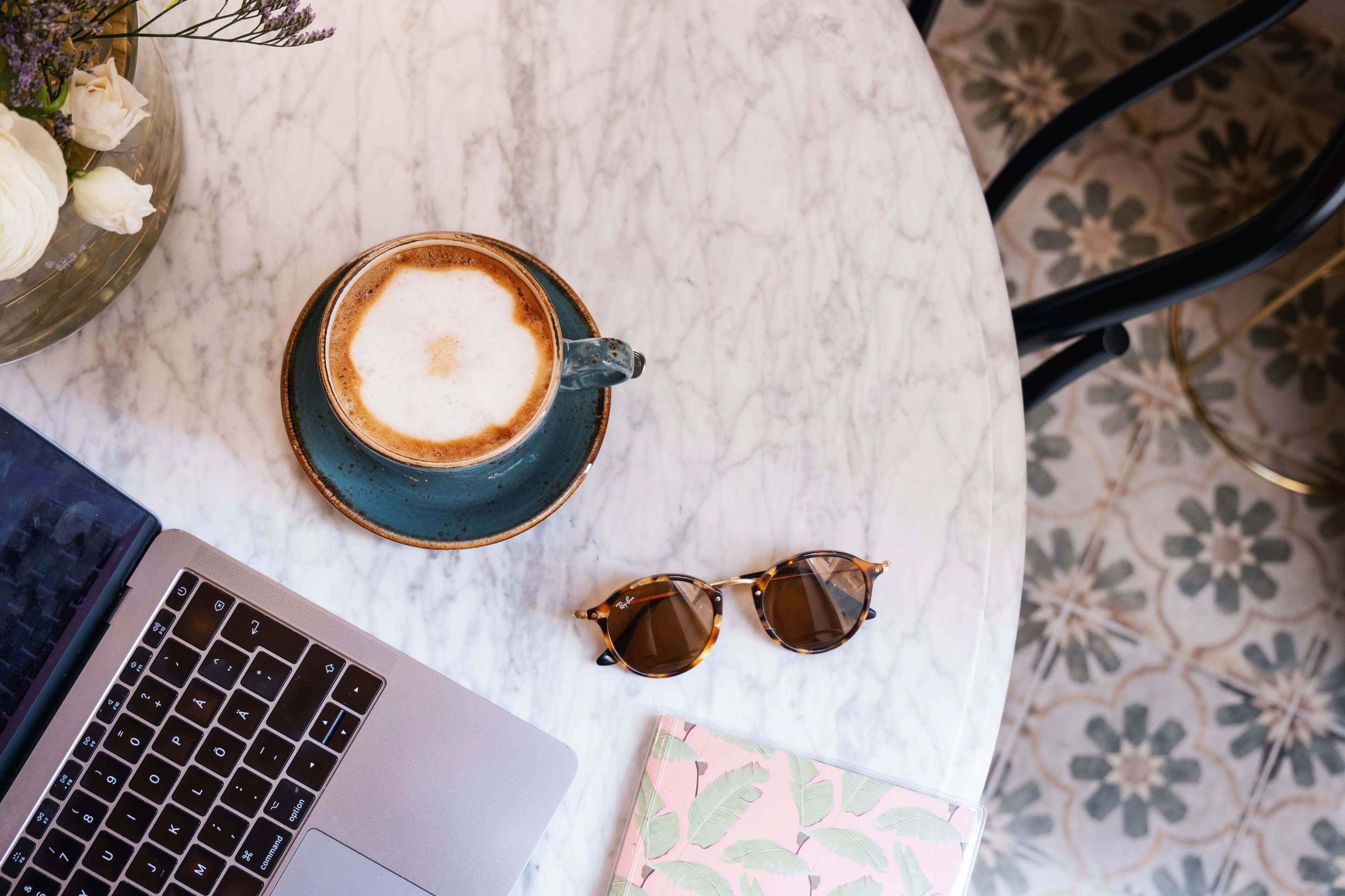 Una laptop, una taza de café, unos lentes de sol y un cuaderno sobre una mesa de mármol.