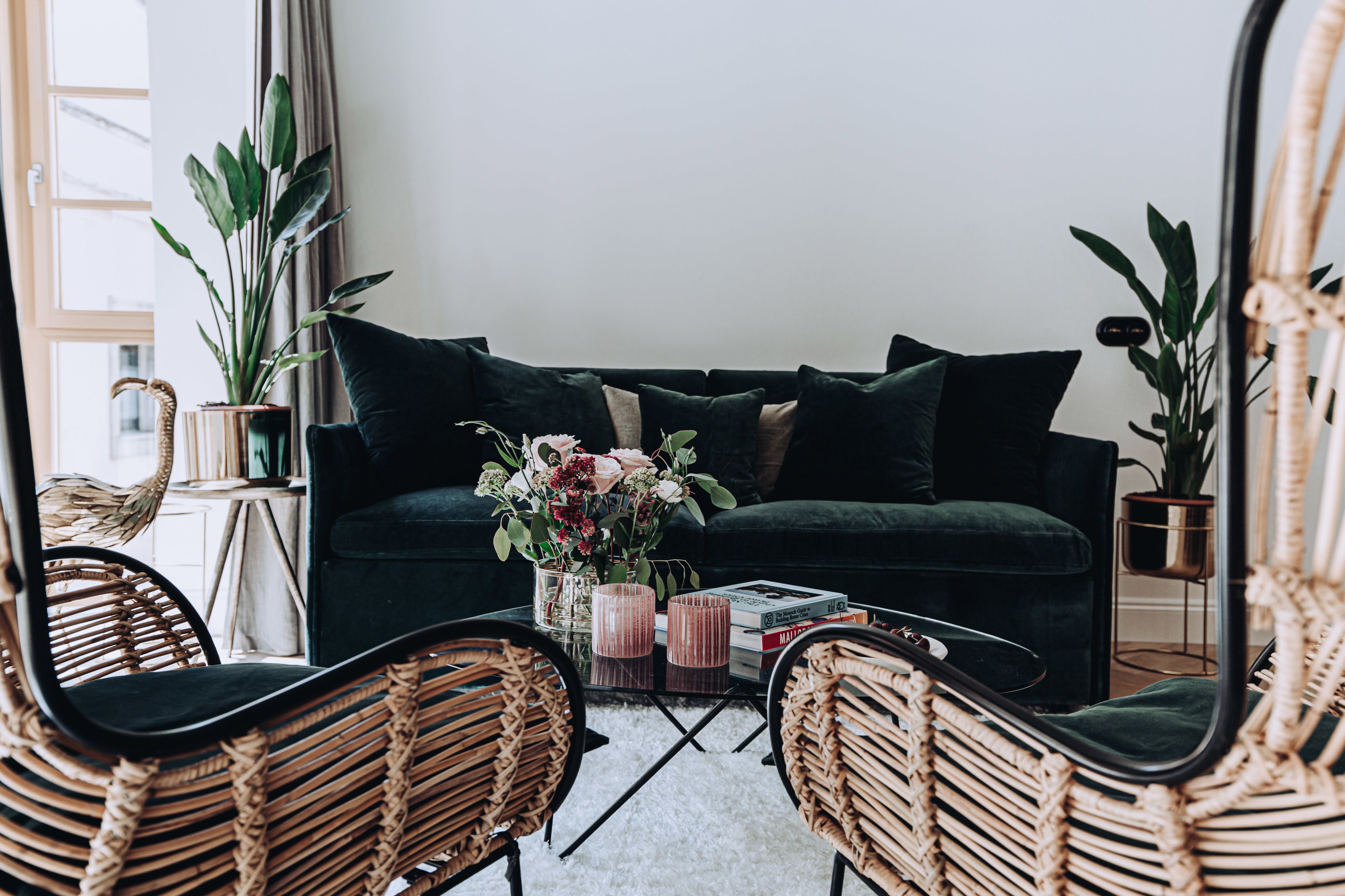 A cozy living room with a dark green sofa, wicker chairs, a coffee table with books and a vase of flowers, and a large mirror reflecting the room.
