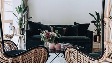 A cozy living room with a dark green sofa, wicker chairs, a coffee table with books and a vase of flowers, and a large mirror reflecting the room.