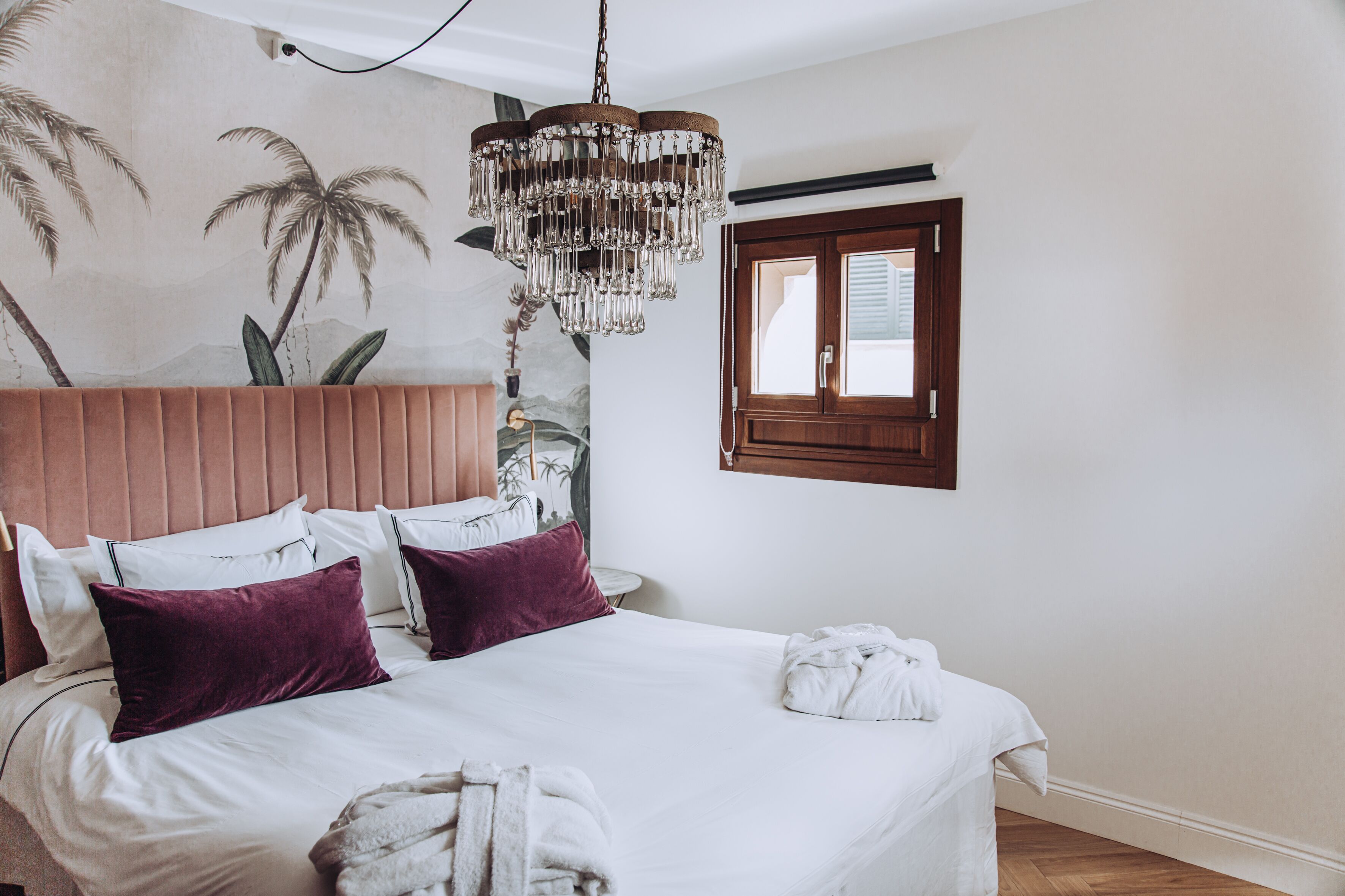 Un dormitorio con cabecera de madera, ropa de cama blanca, una araña y un mural de palmeras.