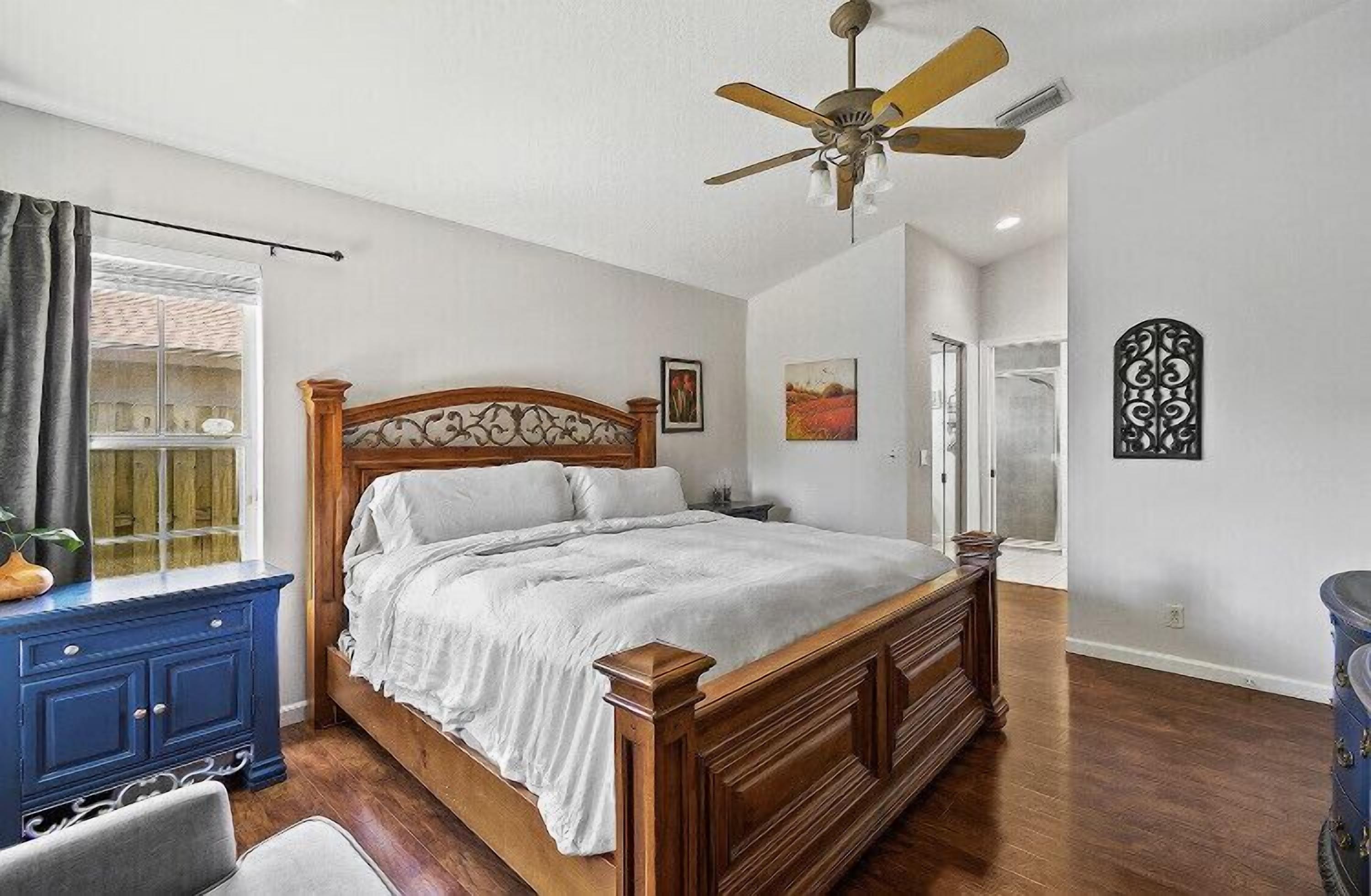 Een slaapkamer met een houten bed, een plafondventilator, een dressoir en een raam met gordijnen.