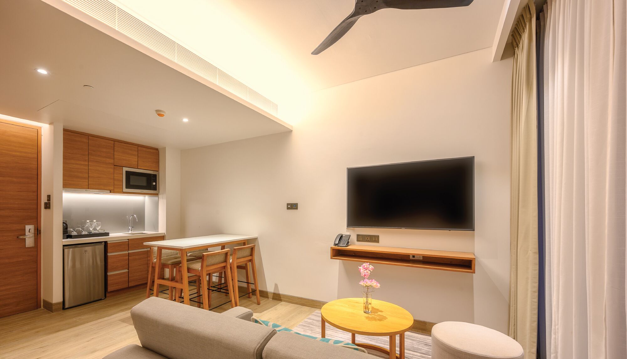 A modern kitchen with wooden cabinets, a microwave, and a sink. There is a dining area with a table and chairs, a TV mounted on the wall, and a small round table with a vase of flowers.