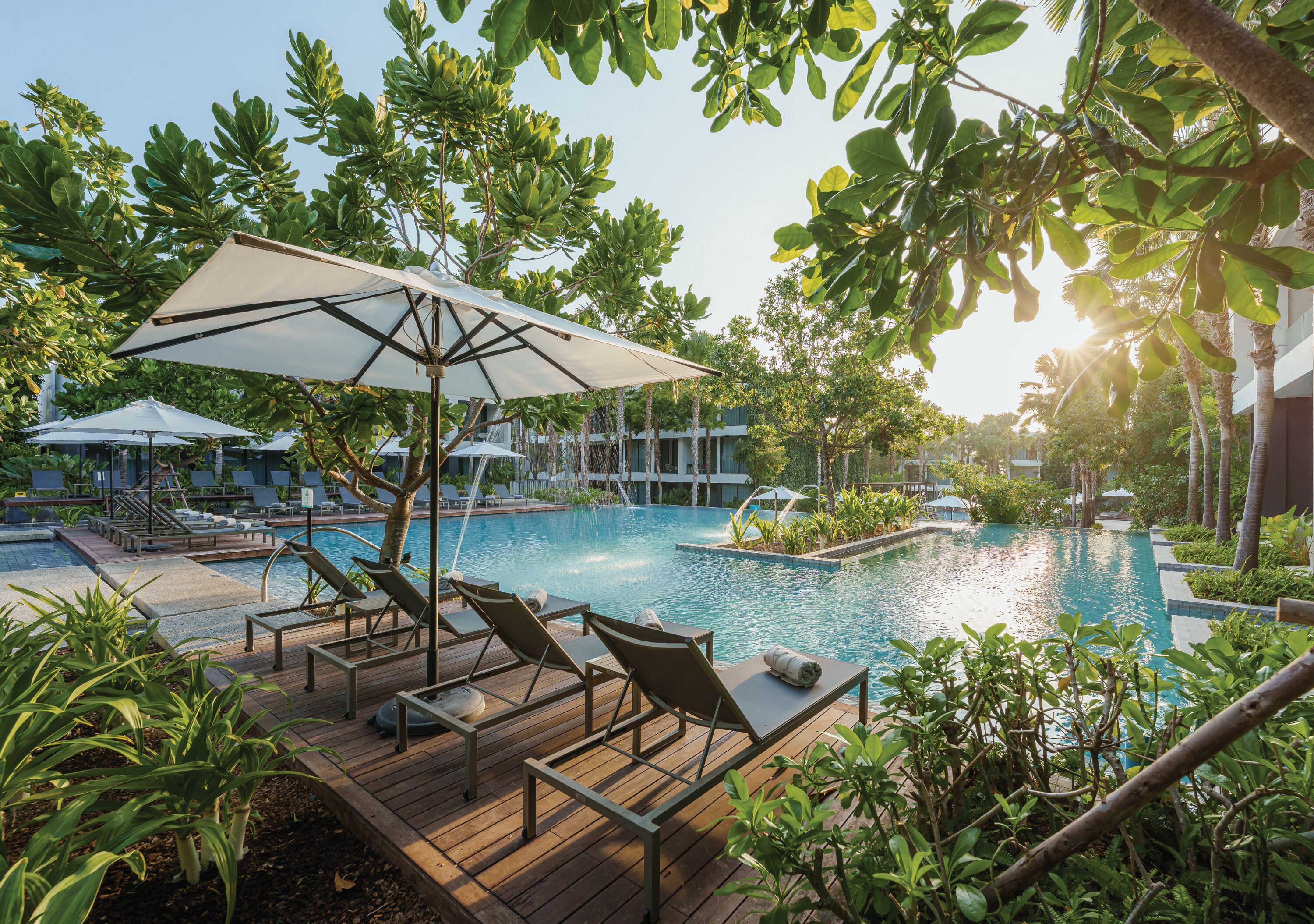 A poolside area with lounge chairs and umbrellas, surrounded by greenery and a clear sky.