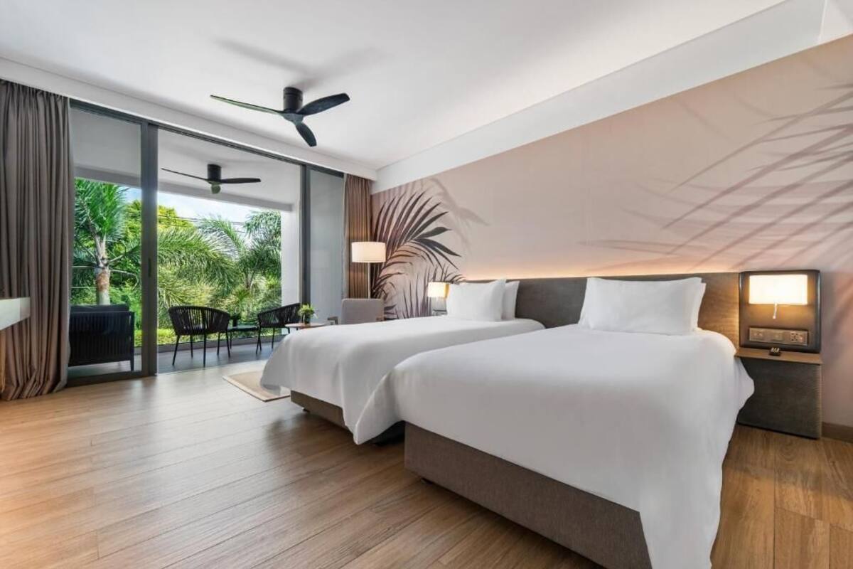 A modern hotel room with two beds, a ceiling fan, a view of greenery, and a dining area with chairs.