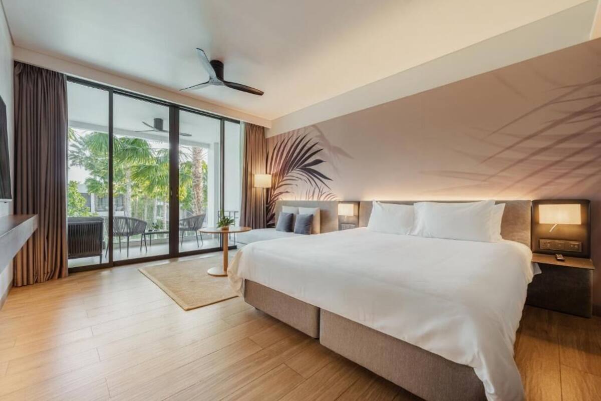 A modern hotel room with a large bed, a ceiling fan, a sliding glass door to the outdoors, and a small table with a lamp.