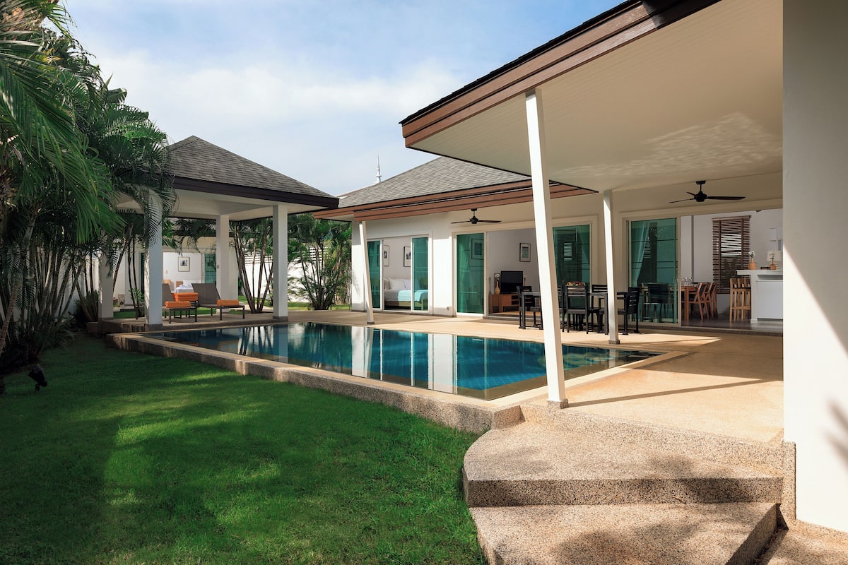 A covered outdoor area with a swimming pool, a patio with seating, and a view of a house in the background.