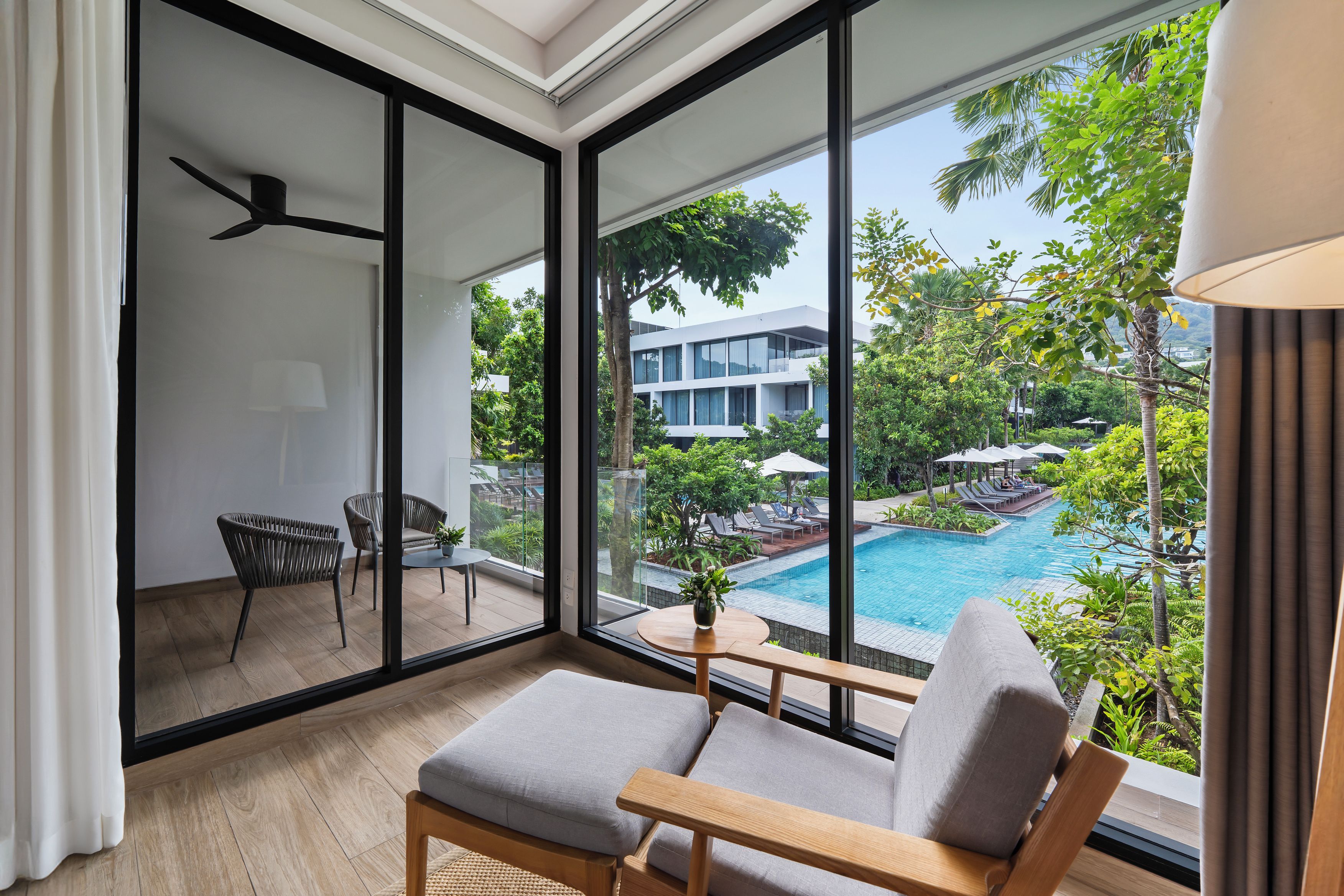 A modern hotel room with a large sliding glass door leading to a pool area, a comfortable seating area with a view, and a ceiling fan.