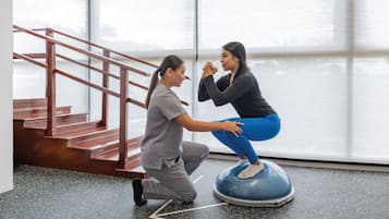 Una persona ayudando a otra en una plataforma de equilibrio en una habitación con un ventanal y una escalera.
