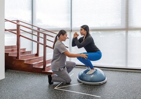Una persona ayudando a otra en una plataforma de equilibrio en una habitación con un ventanal y una escalera.