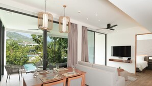 A modern hotel room with a dining area, a sofa, a TV, and a balcony with a pool view. - Stay Wellbeing & Lifestyle Resort (Rawai)