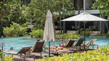 Un espace aménagé au bord de la piscine, avec des chaises longues et des parasols, entouré d’une végétation luxuriante et avec un bâtiment moderne en arrière-plan.