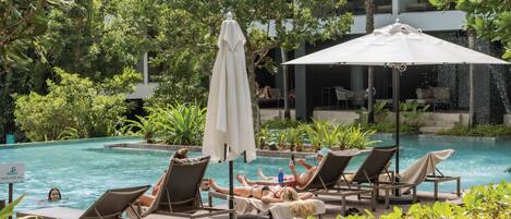 A poolside area with lounge chairs and umbrellas, surrounded by lush greenery and a modern building in the background.