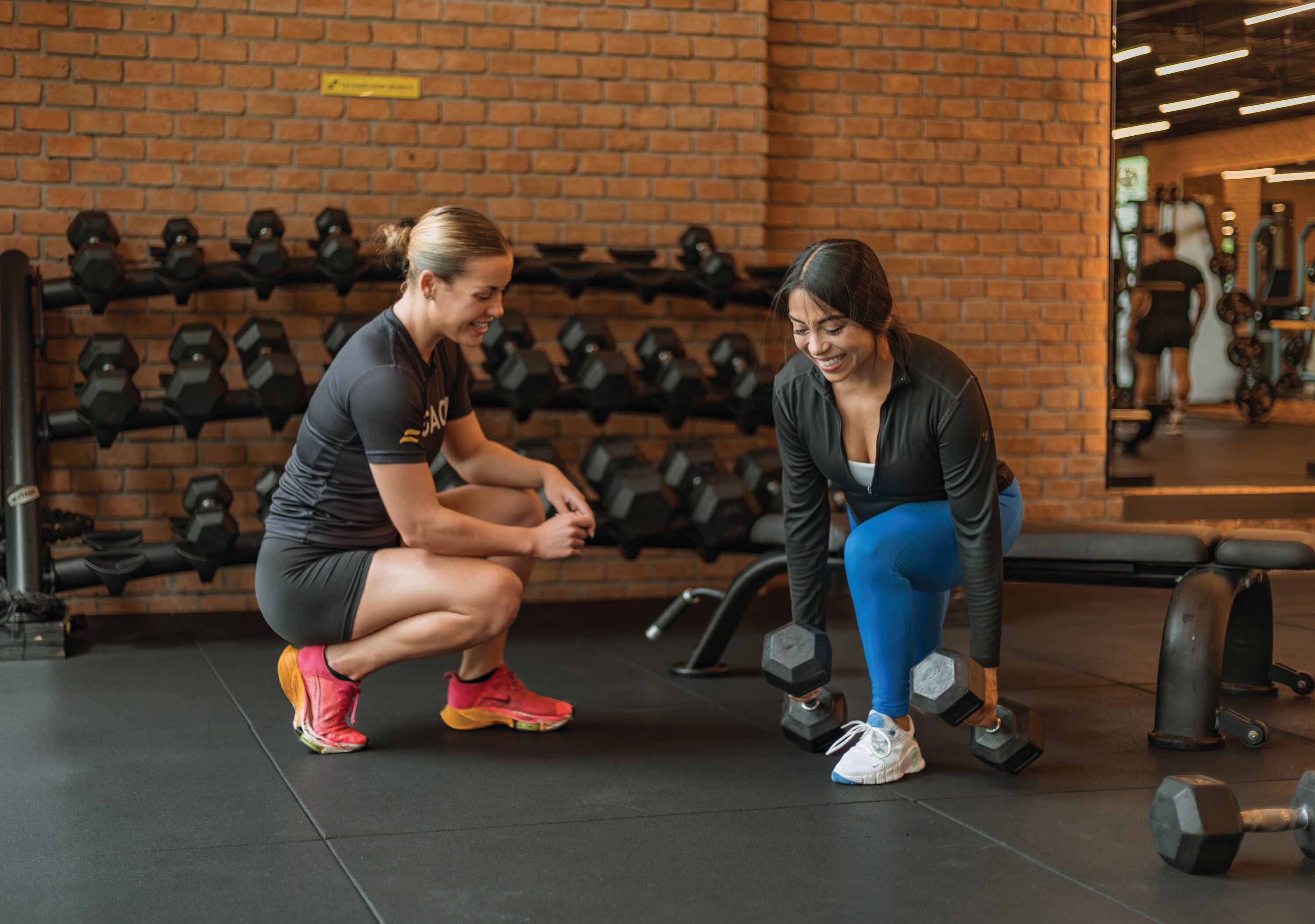 Zwei Personen im Fitnessstudio mit Kurzhanteln und einer Ziegelwand.