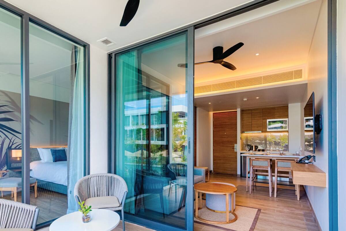A modern hotel room with a glass wall, a ceiling fan, a dining area with a wooden table and chairs, and a cozy seating area with a sofa and armchairs.