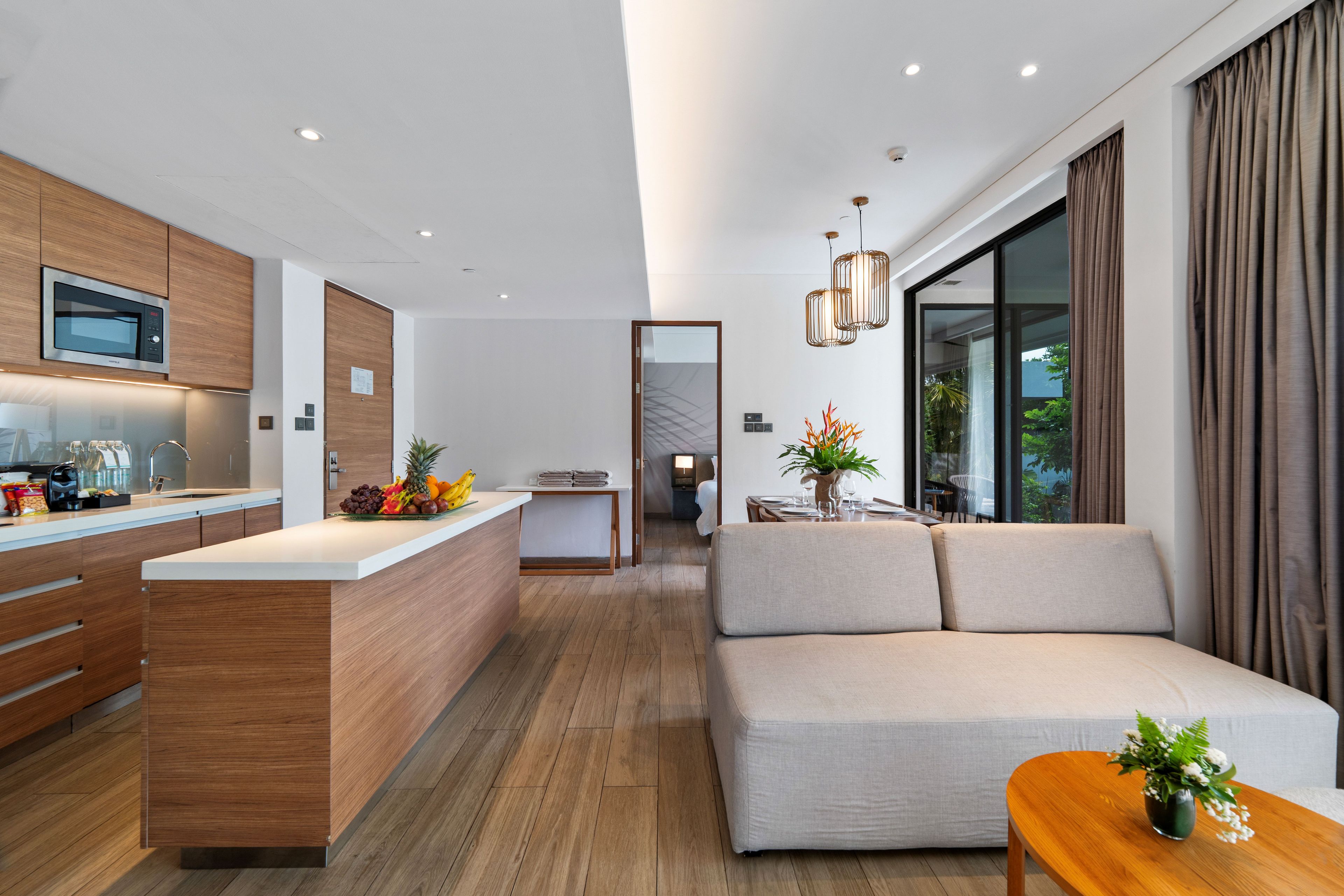Una cocina moderna con microondas, frutero y mueble de madera. Hay un sofá y una mesa de comedor con un jarrón de flores.