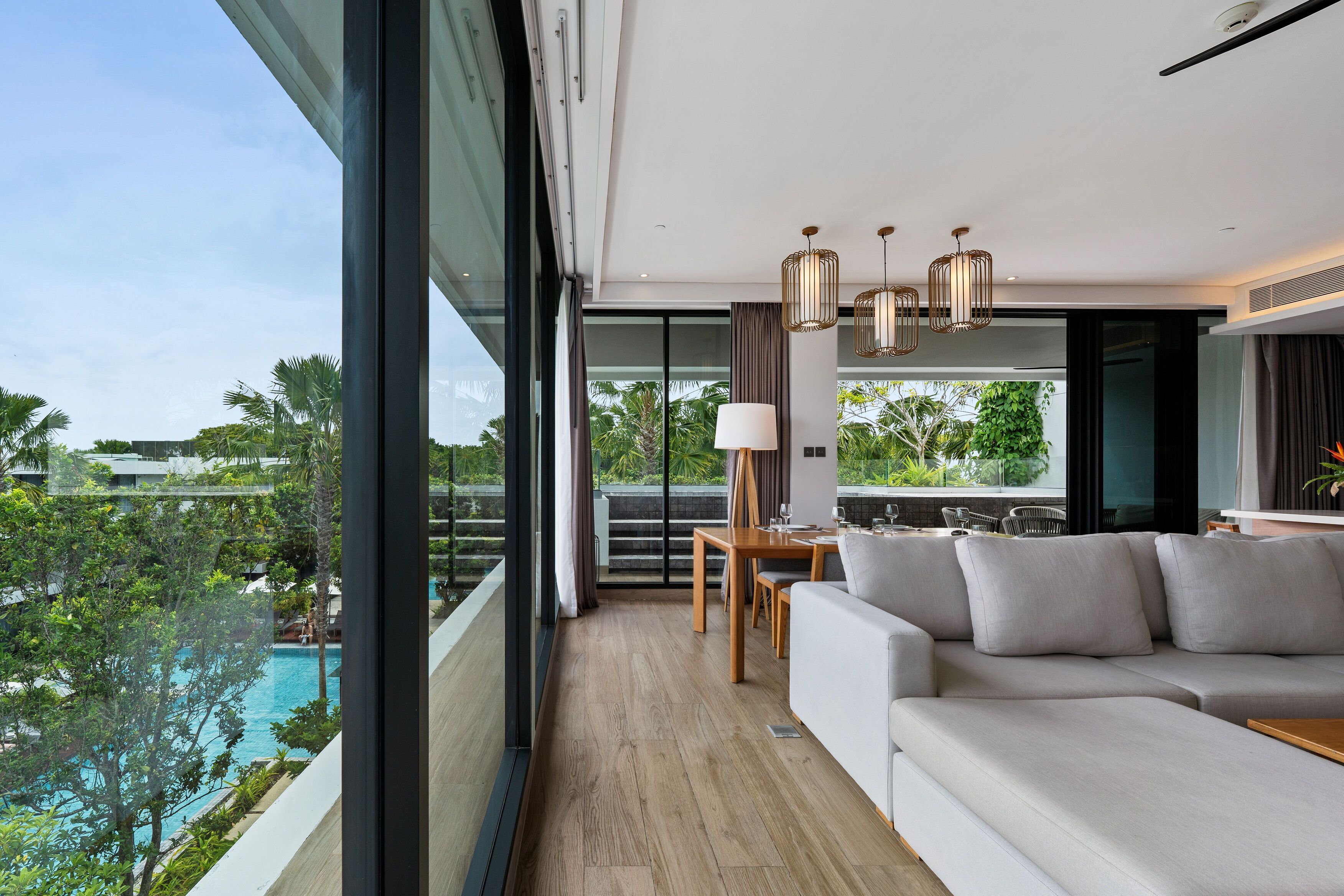 A modern living room with a large window, a sofa, a dining table, and a view of a pool and greenery.