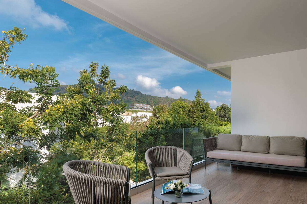 A balcony with a sofa and chairs, a table with a magazine and flowers, and a view of trees and buildings.