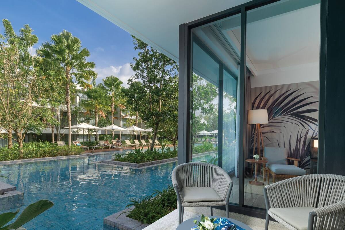 A modern hotel room with a large glass window overlooking a swimming pool and lush greenery. There are two wicker chairs and a small table with a lamp.