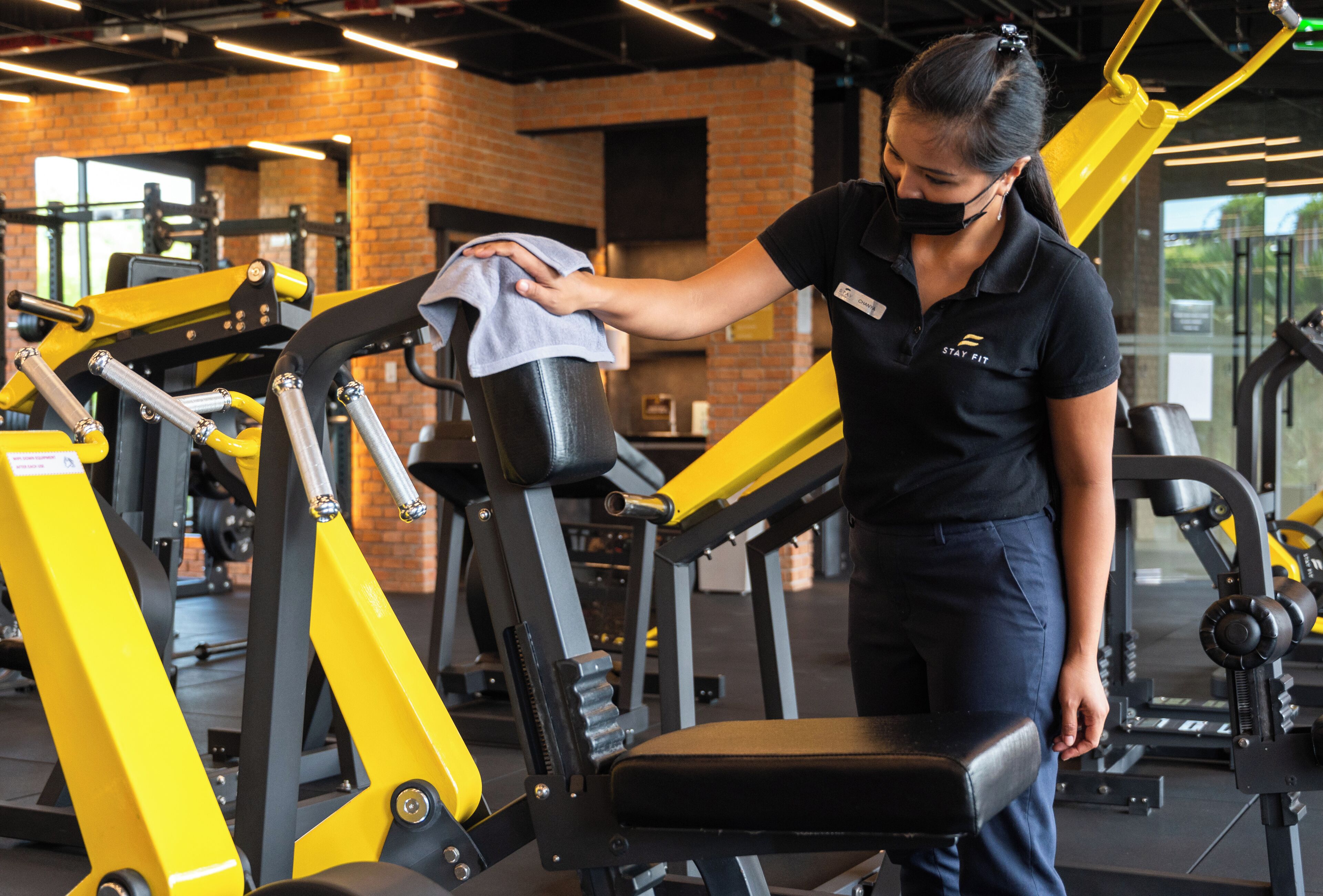 Ein Fitnessstudio mit Trainingsgeräten, eine Person reinigt eine Maschine, und im Hintergrund eine Backsteinwand.