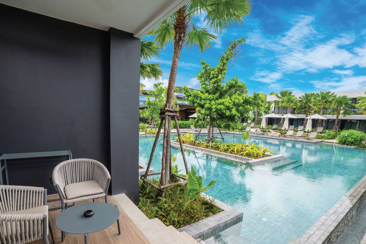 A modern pool area with lounge chairs and a small table, surrounded by lush greenery and a clear blue sky.