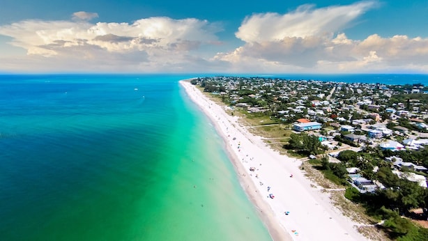 An aerial view of a coastal area with a long sandy beach, clear turquoise waters, and a densely populated residential area with houses and buildings. - A short walk to the beach and direct pool access! (Bradenton Beach)