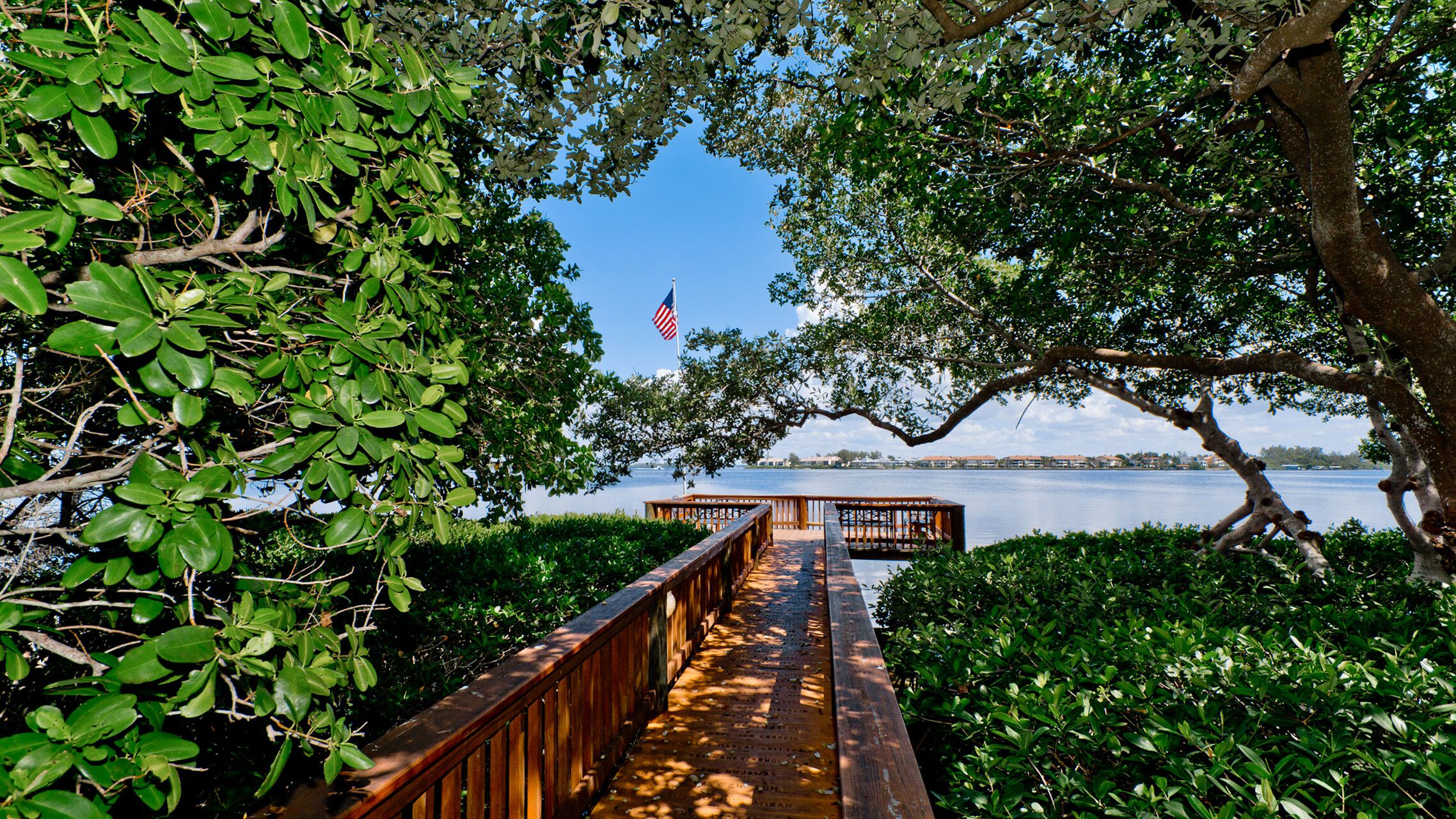 A wooden boardwalk leading to a dock with an American flag, surrounded by lush greenery and a calm body of water.