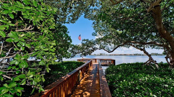 A wooden boardwalk leading to a dock with an American flag, surrounded by lush greenery and a calm body of water.