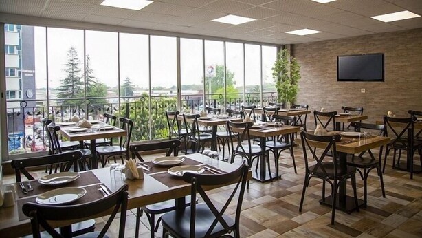 Um restaurante com várias mesas e cadeiras, grandes janelas com vista para a cidade e uma televisão de tela plana na parede.