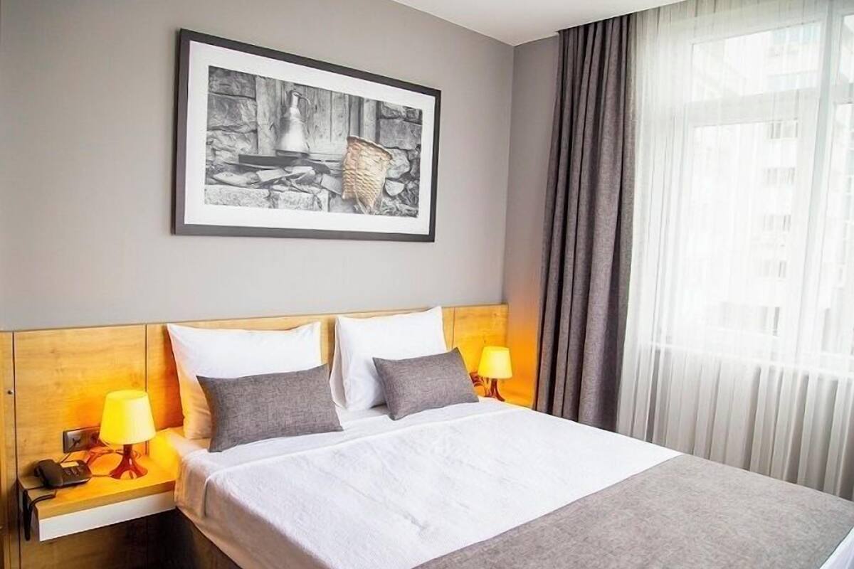 A hotel room with a bed, two bedside lamps, a framed picture on the wall, and a window with curtains.