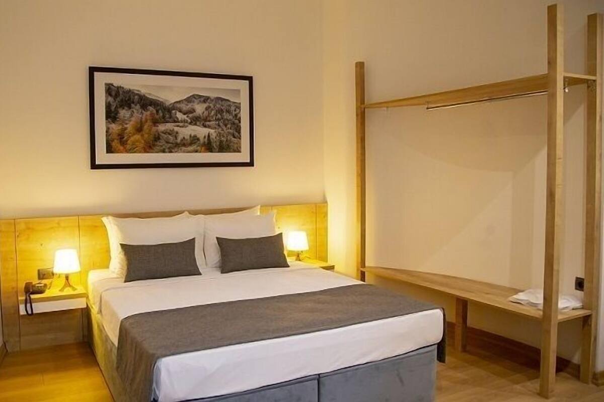 A hotel room with a bed, two bedside lamps, a wooden headboard, a wooden bench, and a framed landscape painting on the wall.
