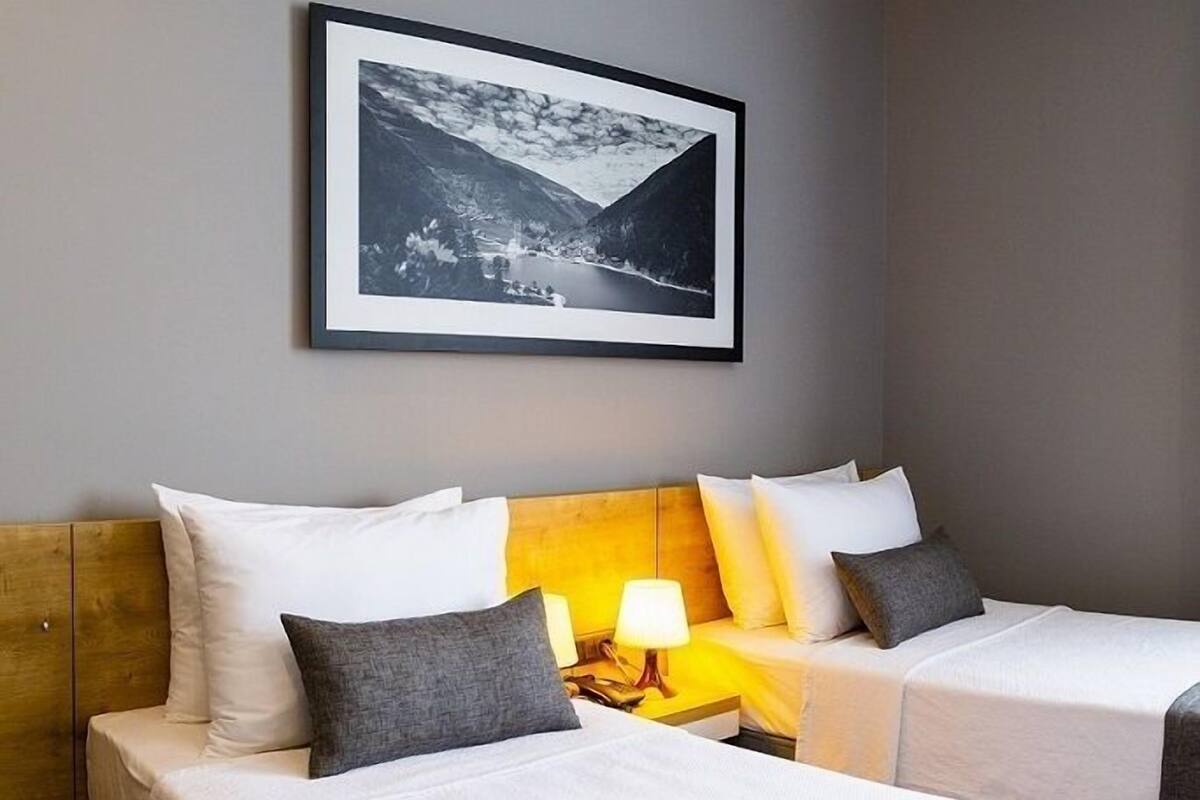 A hotel room with two beds, a wooden headboard, white bedding, a framed photograph on the wall, and a bedside lamp.