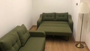Quarto compacto com um sofá verde e um sofá menor, uma luminária de chão e piso de madeira.