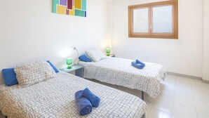 Une chambre d’hôtel avec deux lits, chacun recouvert d’une couvre-lit à motifs et agrémenté de coussins bleus. Un tableau coloré orne le mur, et une fenêtre aux cadres en bois laisse entrer la lumière.