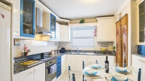 Une cuisine compacte avec des meubles blancs et bleus, un four encastré, un évier et un coin repas avec une table dressée pour deux personnes.