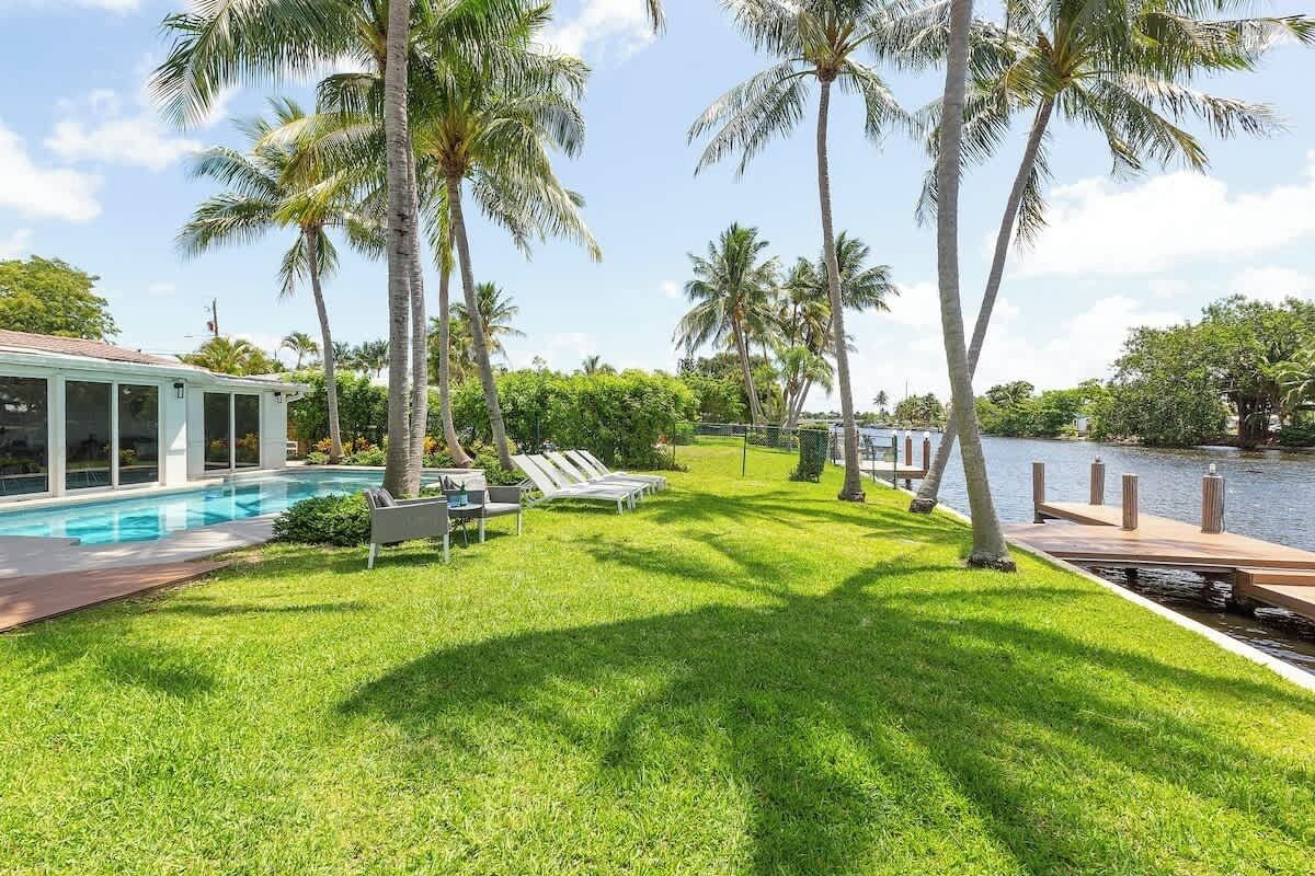 A poolside area with a grassy lawn, lounge chairs, and a wooden dock extending into a body of water, surrounded by palm trees.