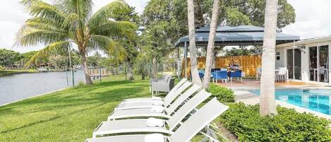 Zona de piscina con sillas de descanso, un área de estar cubierta y vista a un lago.