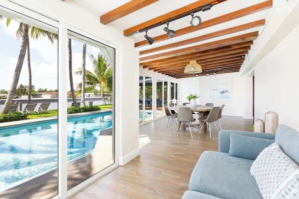 A modern living room with a pool view, wooden ceiling beams, and a dining area.