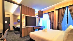 Habitación de hotel con una cama grande, un escritorio con televisor de pantalla plana, una silla y vistas a la ciudad desde las ventanas.