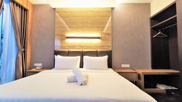 Uma cama bem arrumada com roupa de cama branca e uma toalha enrolada, um telefone de mesa e um luminário fixado na parede.