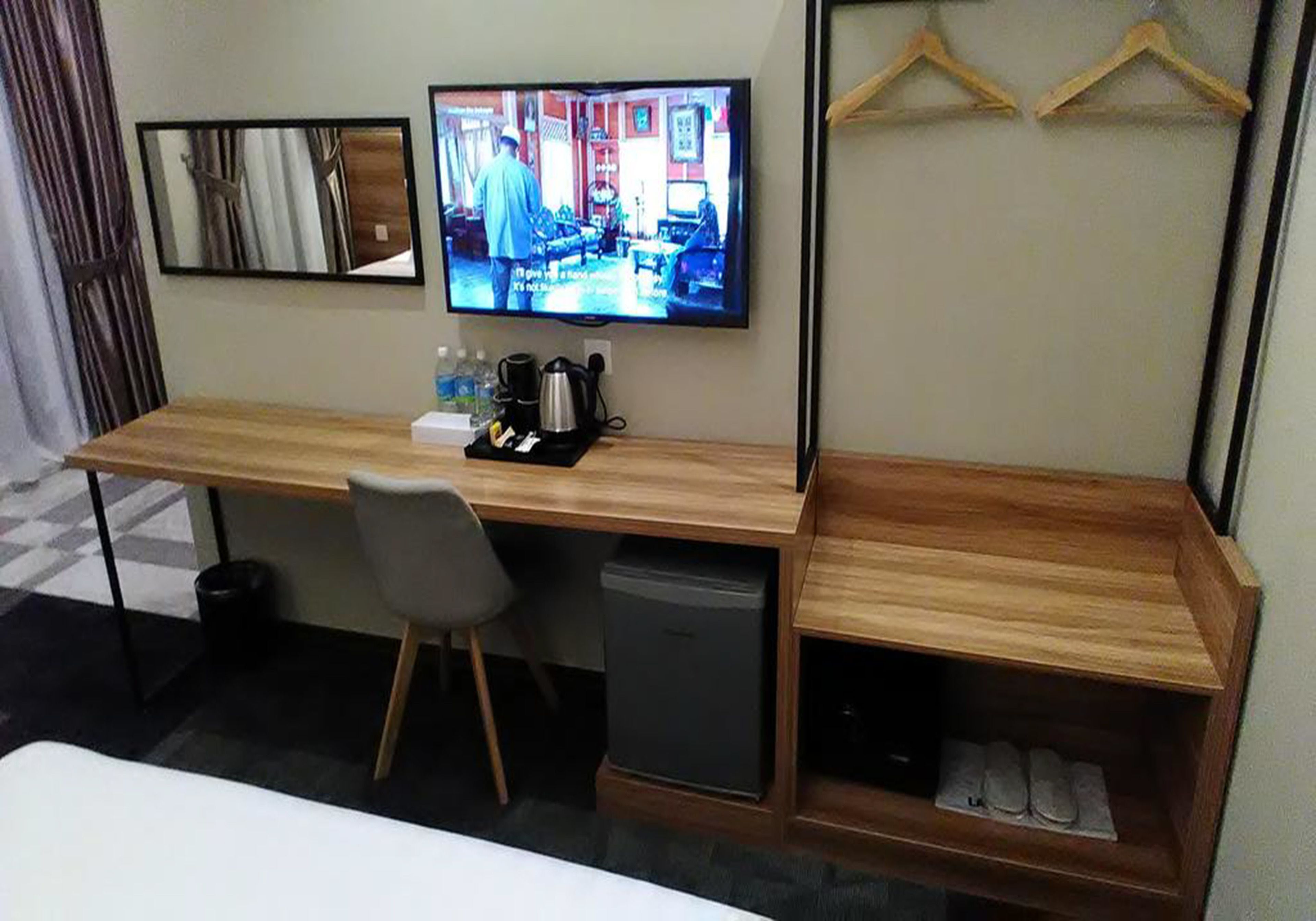 Quarto de hotel com uma mesa de madeira, televisão na parede, lixeira pequena, espaço para guardar sapatos e uma cadeira.
