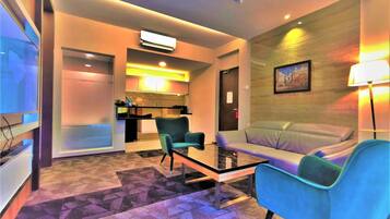 A modern hotel room with a sofa, a small table, and a TV. There is a kitchenette in the background and a door leading to another room.