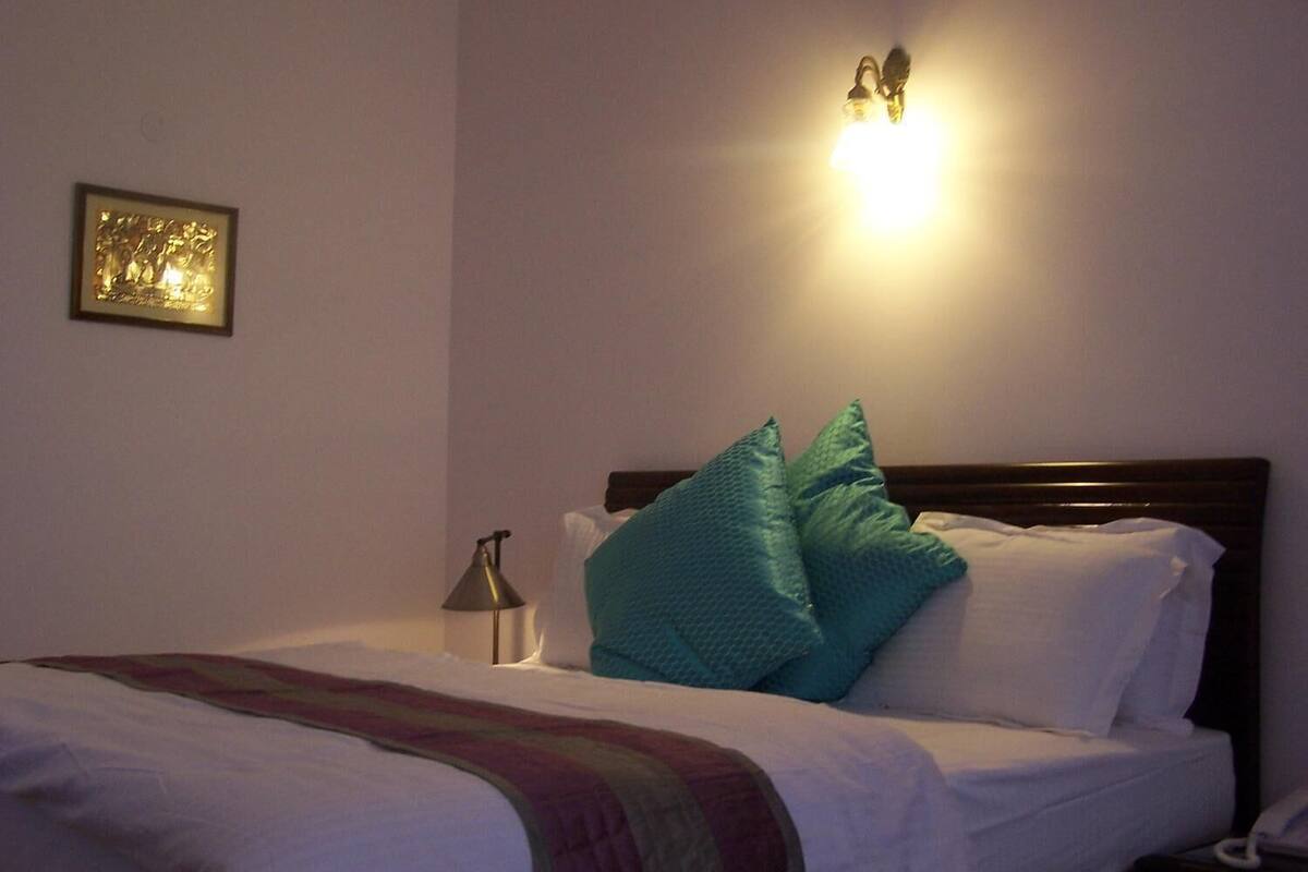A neatly made bed with white linens and teal pillows, a bedside lamp, and a framed picture on the wall.