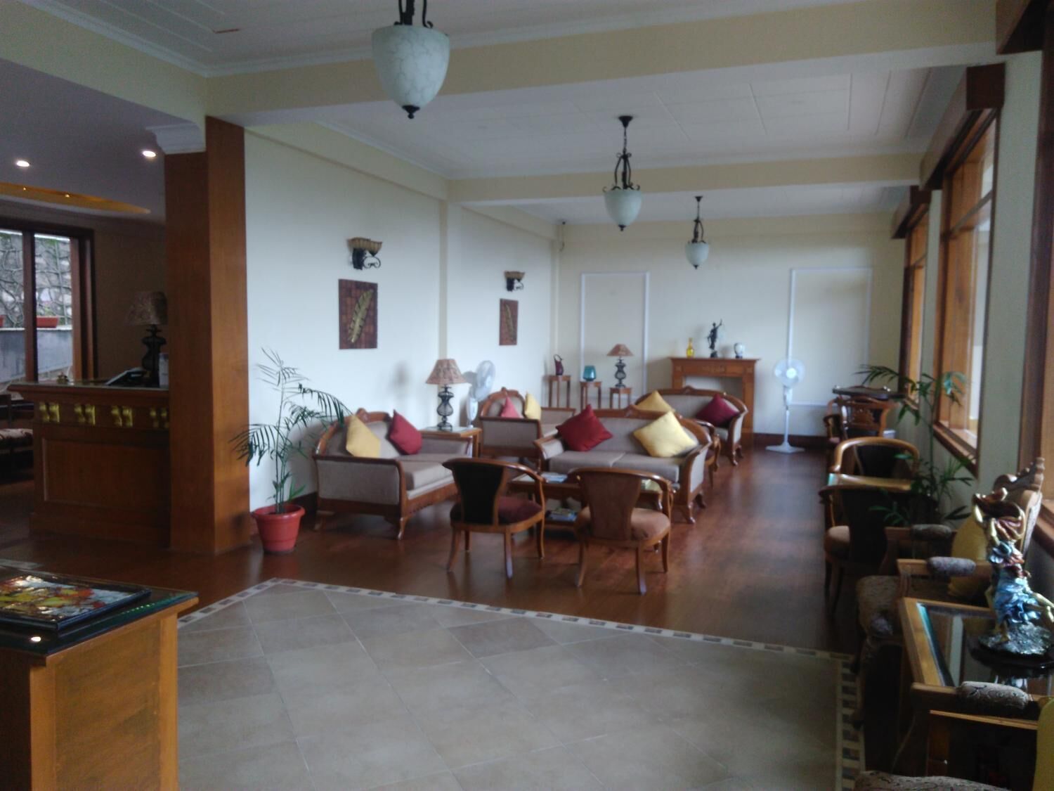 Amplia sala de estar con diversas opciones de asiento, incluyendo sofás y sillones, dispuestos alrededor de una mesa de centro. Hay varios objetos decorativos y plantas, y la habitación cuenta con un alto techo con lámparas colgantes.